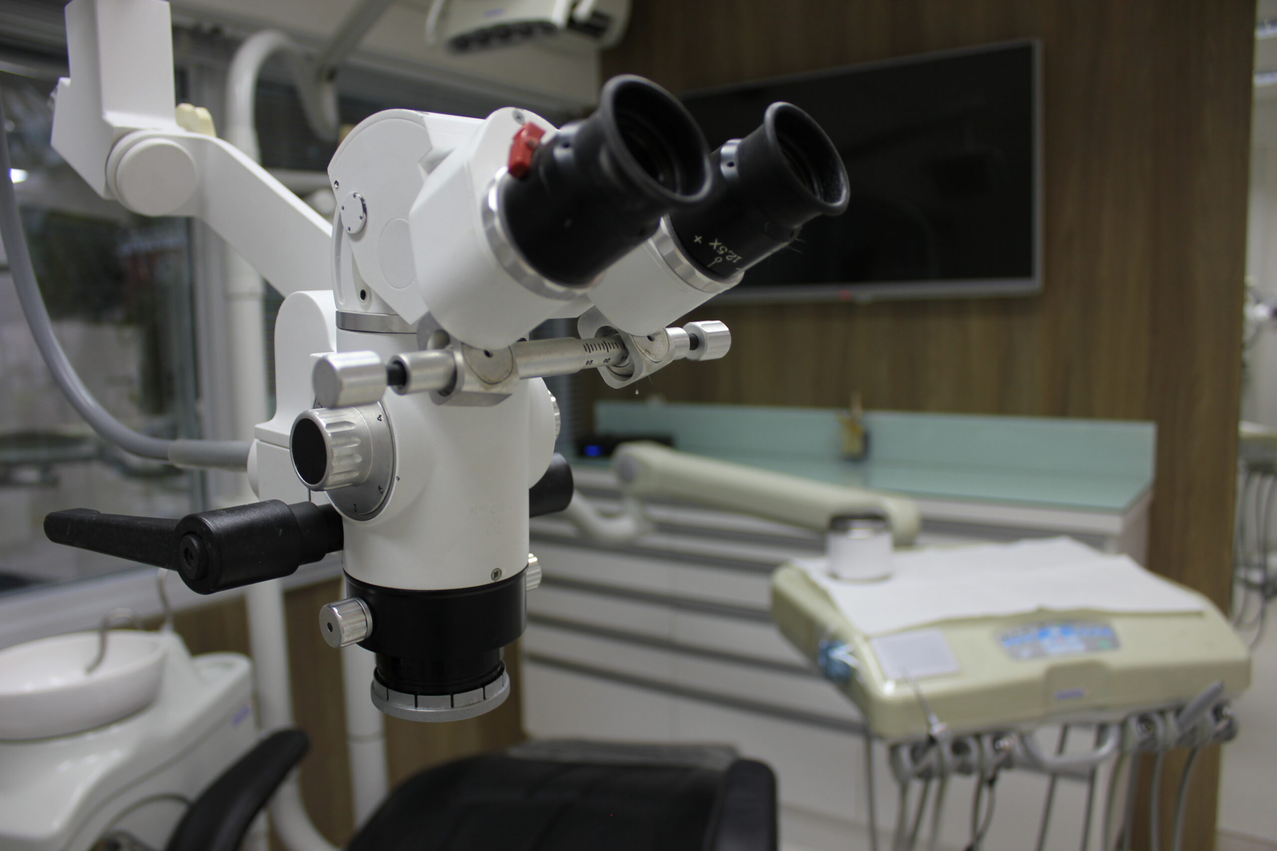 Imagem detalhada de um microscópio odontológico em um consultório moderno. Ao fundo, há uma cadeira odontológica, um monitor para agendamento e equipamentos especializados para o sistema dentista 24 horas, refletindo um ambiente tecnológico e preparado para procedimentos de alta precisão nas consultas e na urgência e emergência.