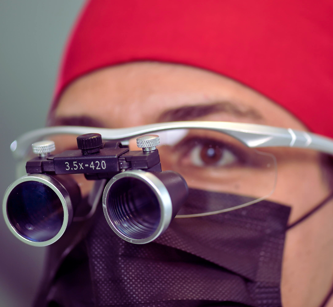 Dentista 24 horas de plantão utilizando máscara, um gorro vermelho e uma lupa de aumento 3,5x, preparado para atendimento de urgência e emergência no sistema dentista 24 horas e agendamento de pacientes.