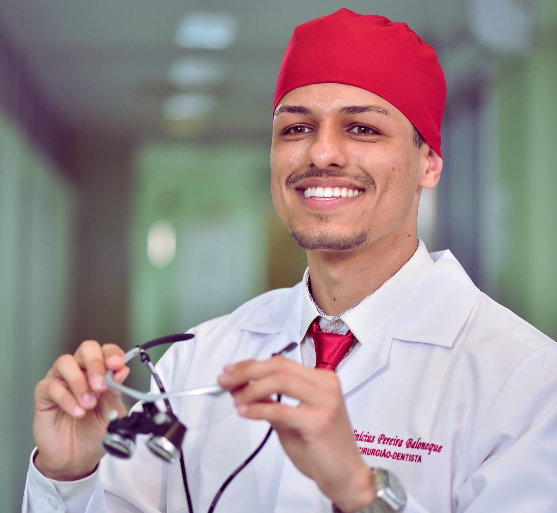 Dentista 24 horas de plantão segurando uma lupa de aumento 3,5x usando jaleco branco, toca e gravata vermelha, passando profissionalismo e confiança para o atendimento de urgência e emergência no sistema dentista 24 horas e agendamento de pacientes.