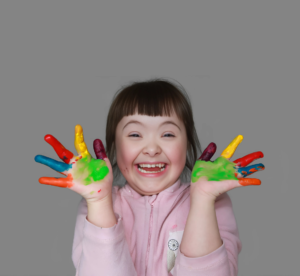 Criança com síndrome de Down sorridente, com as mãos cobertas de tinta colorida, expressando alegria e criatividade. Momento lúdico e divertido, destacando a importância da arte na infância e a inclusão. Mostrando a acessibilidade no sistema de agendamento ou dentista 24 horas.