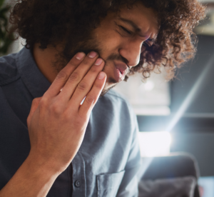 Homem com expressão de dor segurando a lateral do rosto, aparente desconforto devido a dor de dente. Cena ilustrativa de problemas odontológicos, como cáries ou sensibilidade dentária. Possíveis problemas que levam a procura do agendamento ou do sistema dentista 24 horas para atendimento de urgência e emergência.