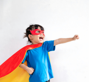 Criança vestida de super-herói, usando uma capa vermelha e amarela, uma máscara e uma camiseta azul, enquanto o dedo voa com o braço estendido. A cena transmite diversão, imaginação e empoderamento.