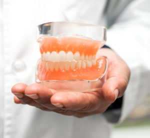 Dentista segurando uma prótese dentária em suas mãos, destacando dentes brancos e gengivas artificiais em um modelo transparente. A imagem simboliza cuidados odontológicos e próteses dentárias, ilustrando os trabalhos oferecidos nas consultas de agendamento ou urgência e emergência.